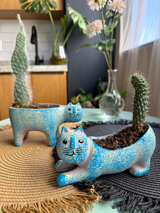 Two blue cat-shaped planters with plants inside on a table.